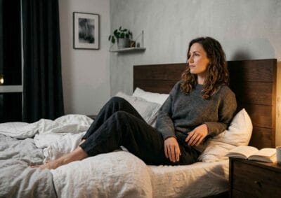 Frau sitzt allein auf einem modernen Bett und blickt nachdenklich zur Seite – die Perspektive der Partnerin, wenn Sexualität unter Druck die Beziehungsdynamik belastet