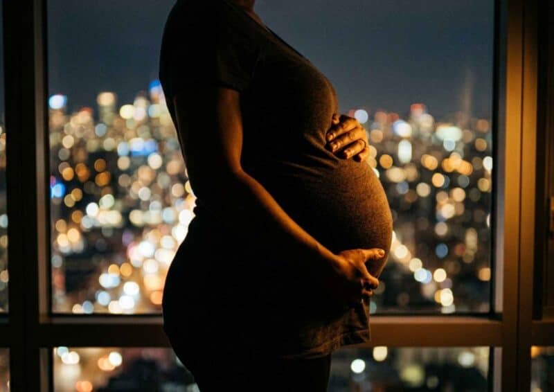 Schwangere Frau vor nächtlicher Stadtsilhouette – Hände auf dem Bauch, warmes Gegenlicht, Stimmung von Stille und innerer Verbundenheit.