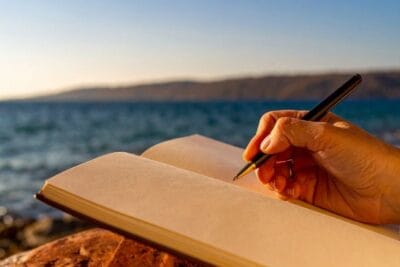 Person sitzt am See und schreibt ihre Wünsche in ein Journalheft für ihr Mood- & Visionboard