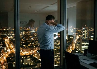 Erschöpfter Mann steht abends am Bürofenster, Hand im Nacken – gestresster Alltag und Nervensystem unter Dauerspannung